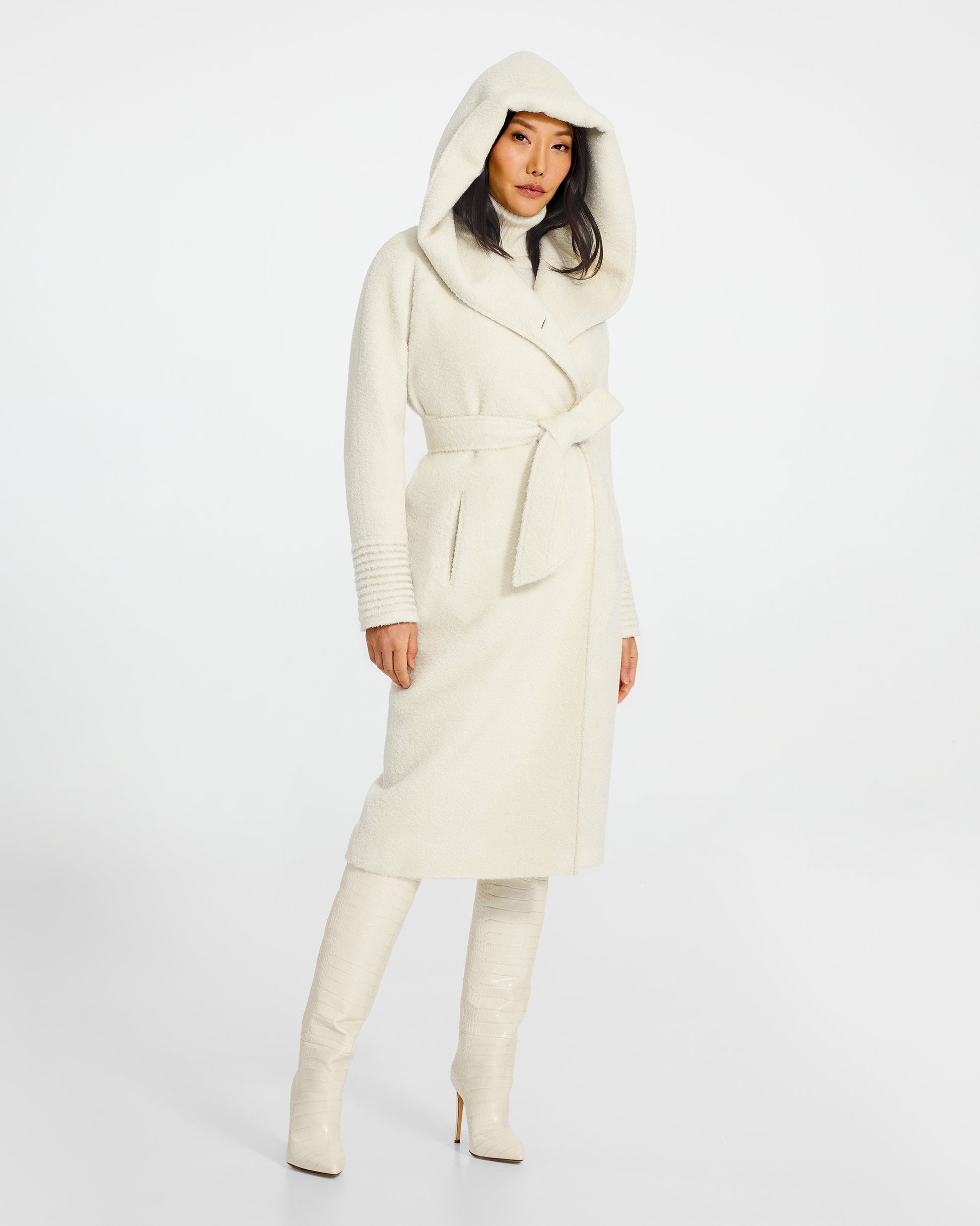 Front view of model wearing a Sentaler long ivory coat made of bouclé alpaca wool, with a hooded collar, worn belted.