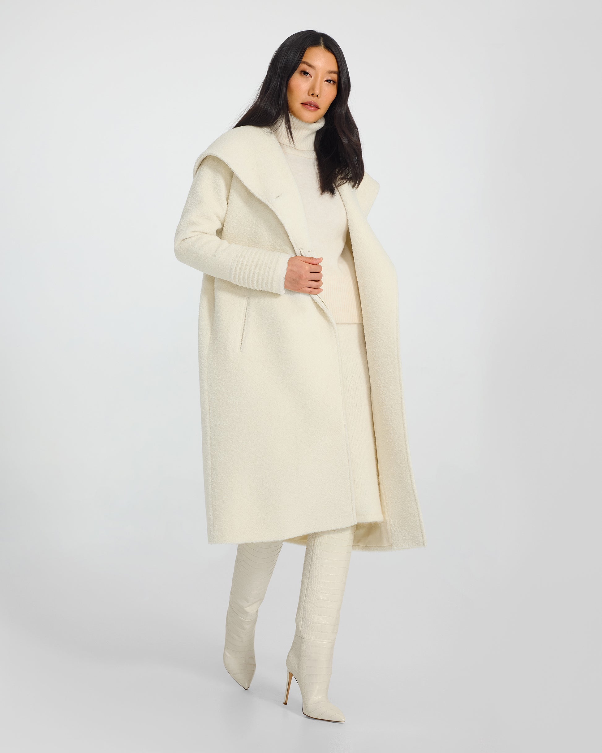 Side view of model wearing a Sentaler long ivory coat made of bouclé alpaca wool, with a hooded collar, worn open.