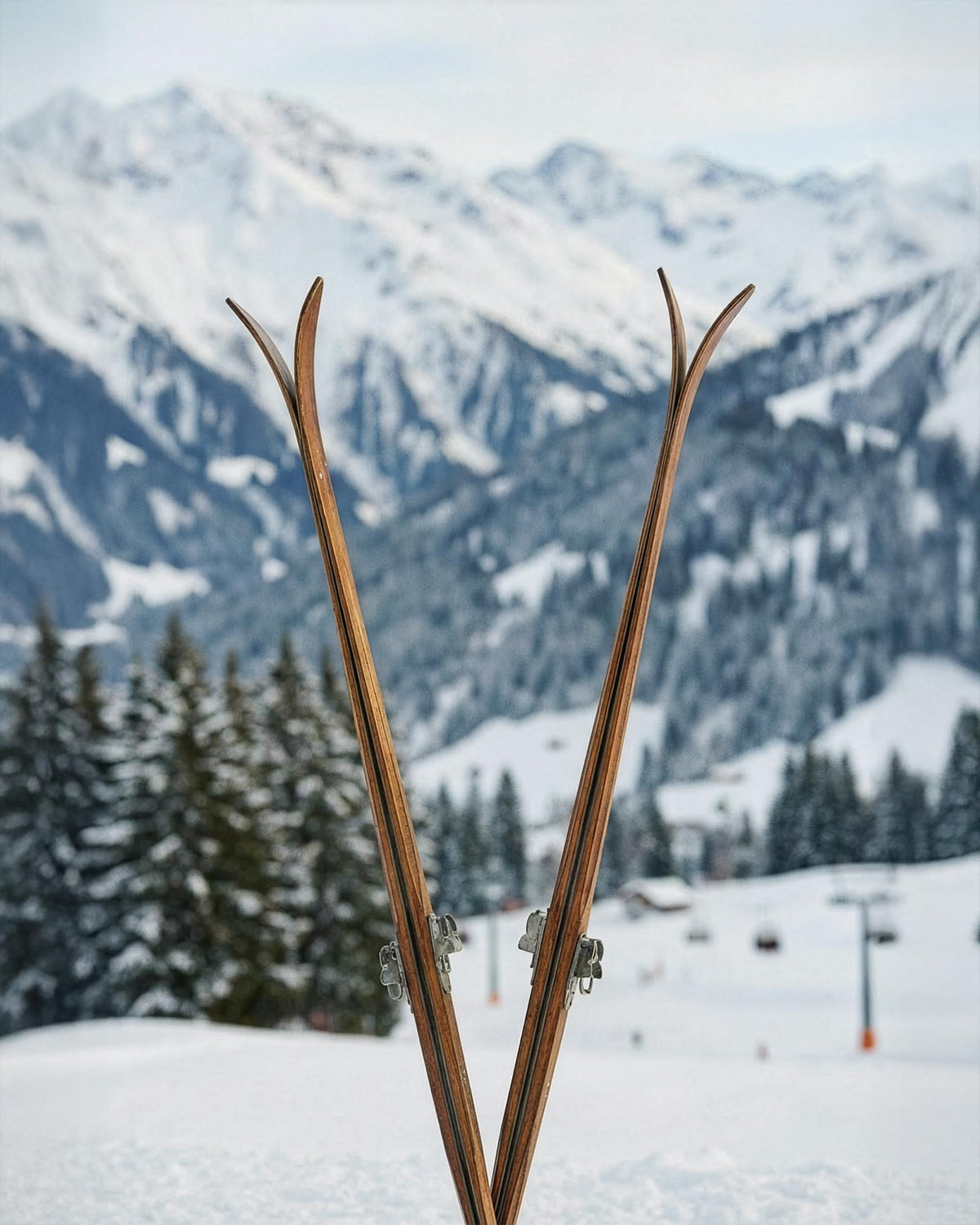 Vintage wooden skis standing upright in the snow with alpine mountains in the background.