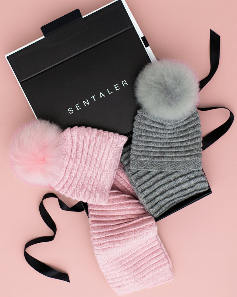 Open SENTALER gift box revealing ribbed knit hats and scarves in blush pink and grey with fur pom-pom details, displayed on a pastel pink background — showcasing cozy luxury gifts for her.