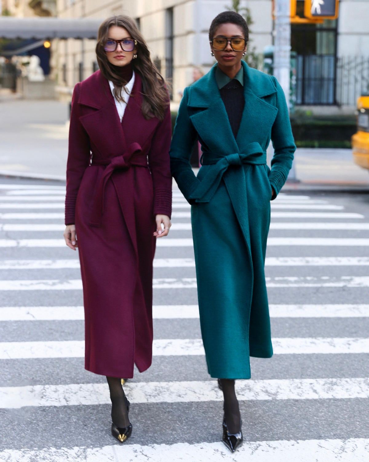 Front view of models wearing Sentaler emerald green & cherry lacquer maxi coats made of suri alpaca wool, with a notched collar, worn belted.