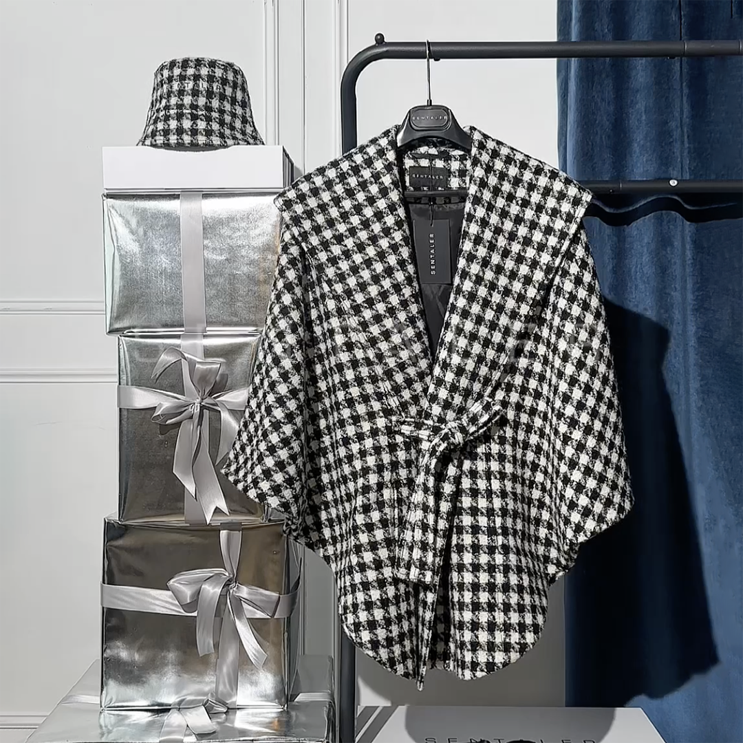 Sentaler Tweed Black and White Houndstooth Cape in Tweed Suri Alpaca wool. Seen hanging on a rack surrounded by wrapped gifts.