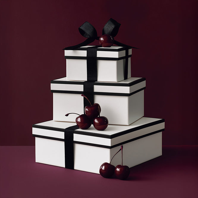 Three white gift boxes wrapped in black ribbon and accented with deep red cherries, styled against a rich burgundy backdrop for the SENTALER Gift Guide.