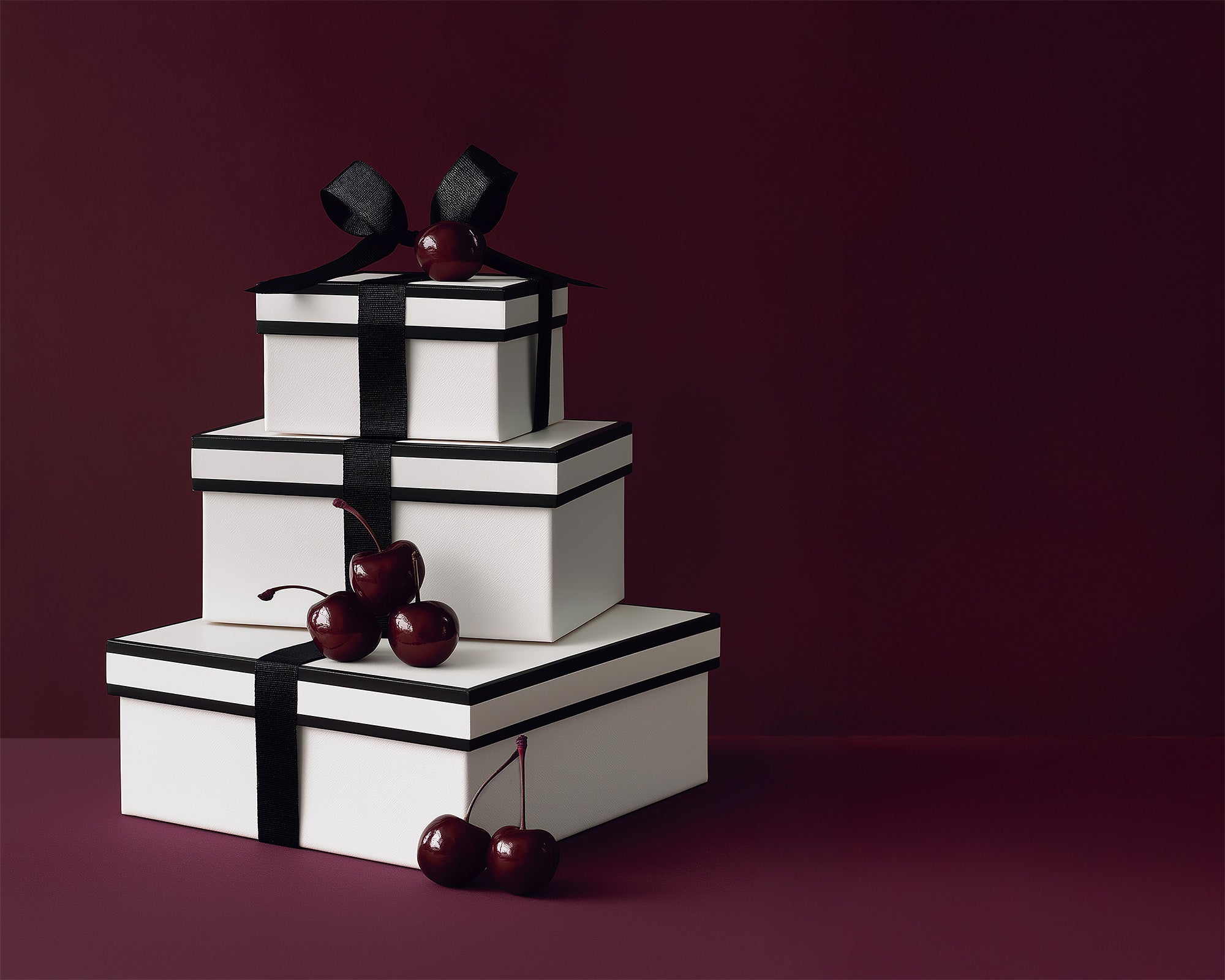Three white gift boxes wrapped in black ribbon and accented with deep red cherries, styled against a rich burgundy backdrop for the SENTALER Gift Guide.