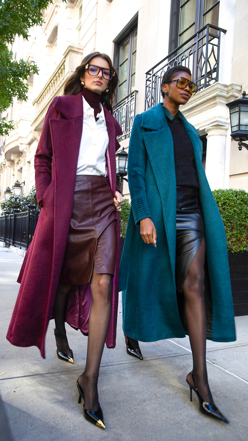 Front close up view of models wearing Sentaler cherry lacquer and emerald maxi coats made of suri alpaca wool, with a notched collar, worn belted.