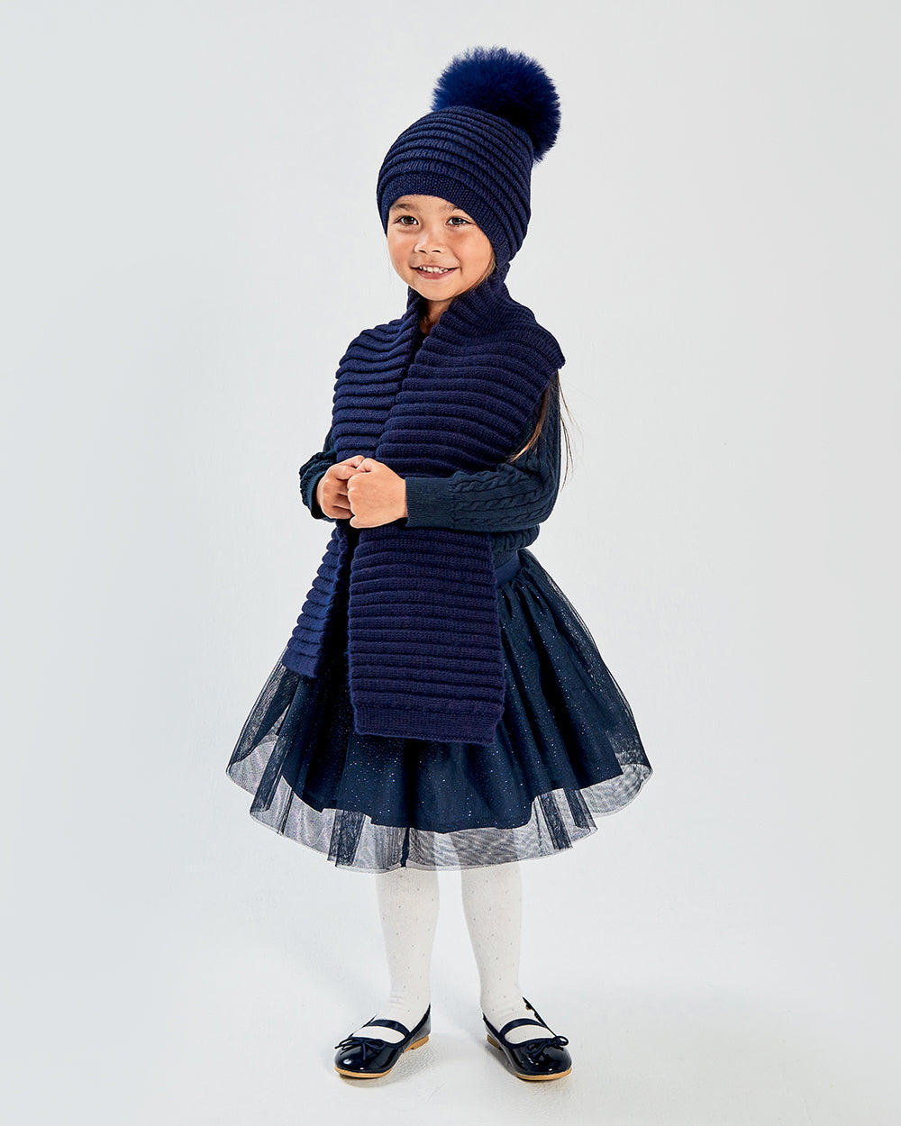 Sentaler Kids Ribbed Navy Hat with Fur Pompon (4-5 Years) and Kids Ribbed Navy Scarf (4-5 Years) in Baby Alpaca yarn. Seen from front on girl model.