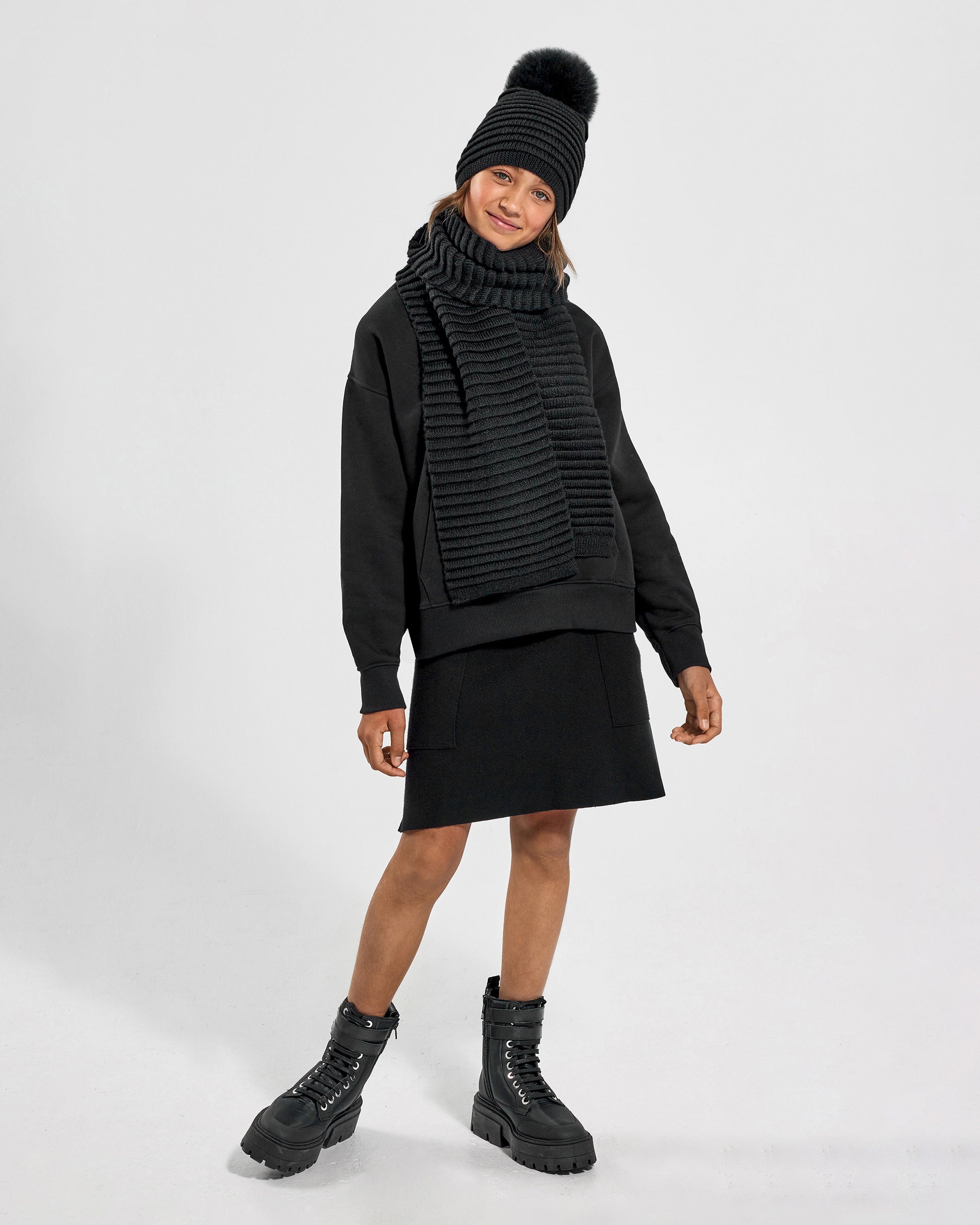 Sentaler Kids Ribbed Black Hat with Fur Pompon (6-14 Years) and Kids Ribbed Black Scarf (6-14 Years) in Baby Alpaca yarn. Seen from front on female model.
