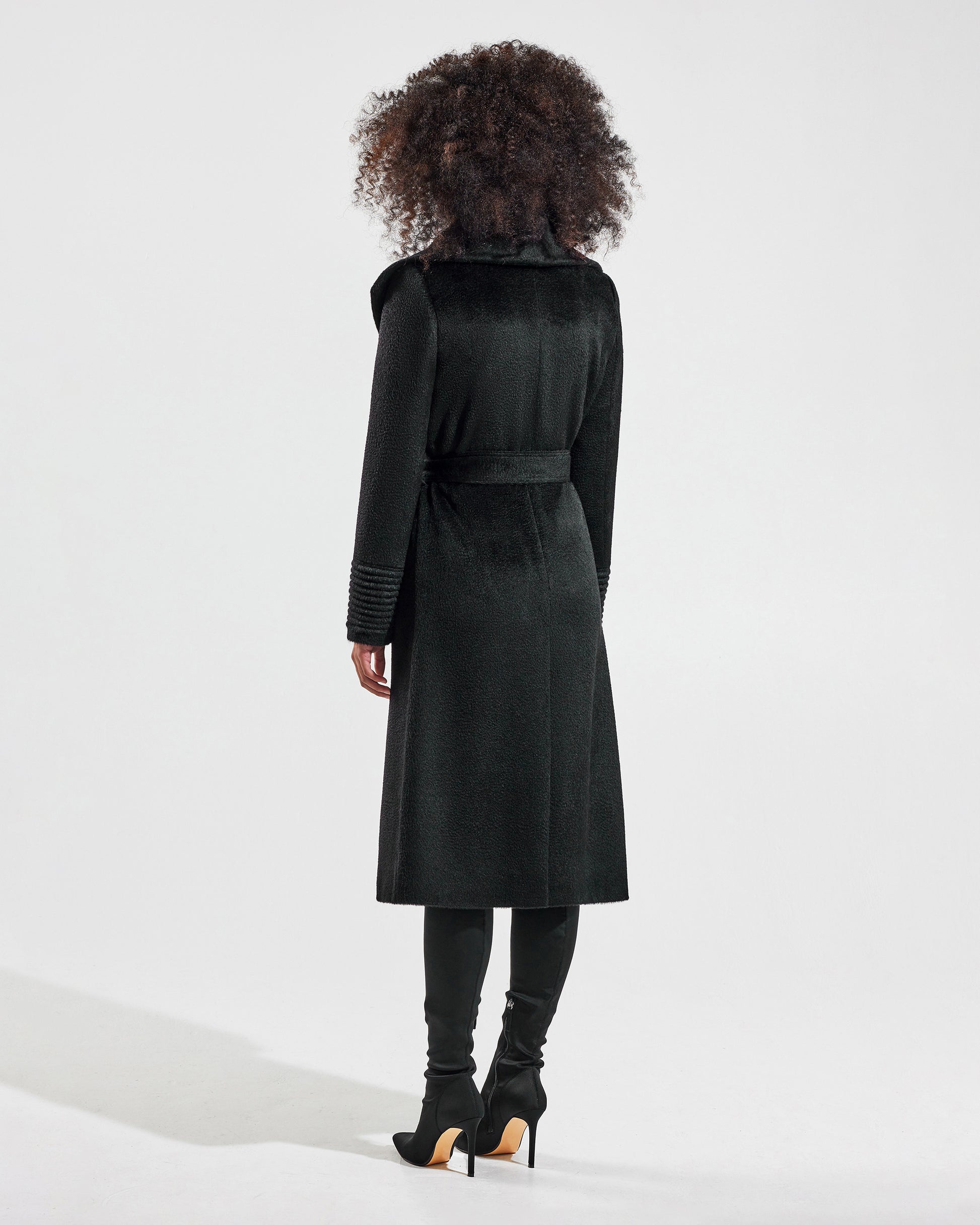 Back view of model wearing a Sentaler long black wrap coat made of suri alpaca wool, with a shawl collar, worn belted.
