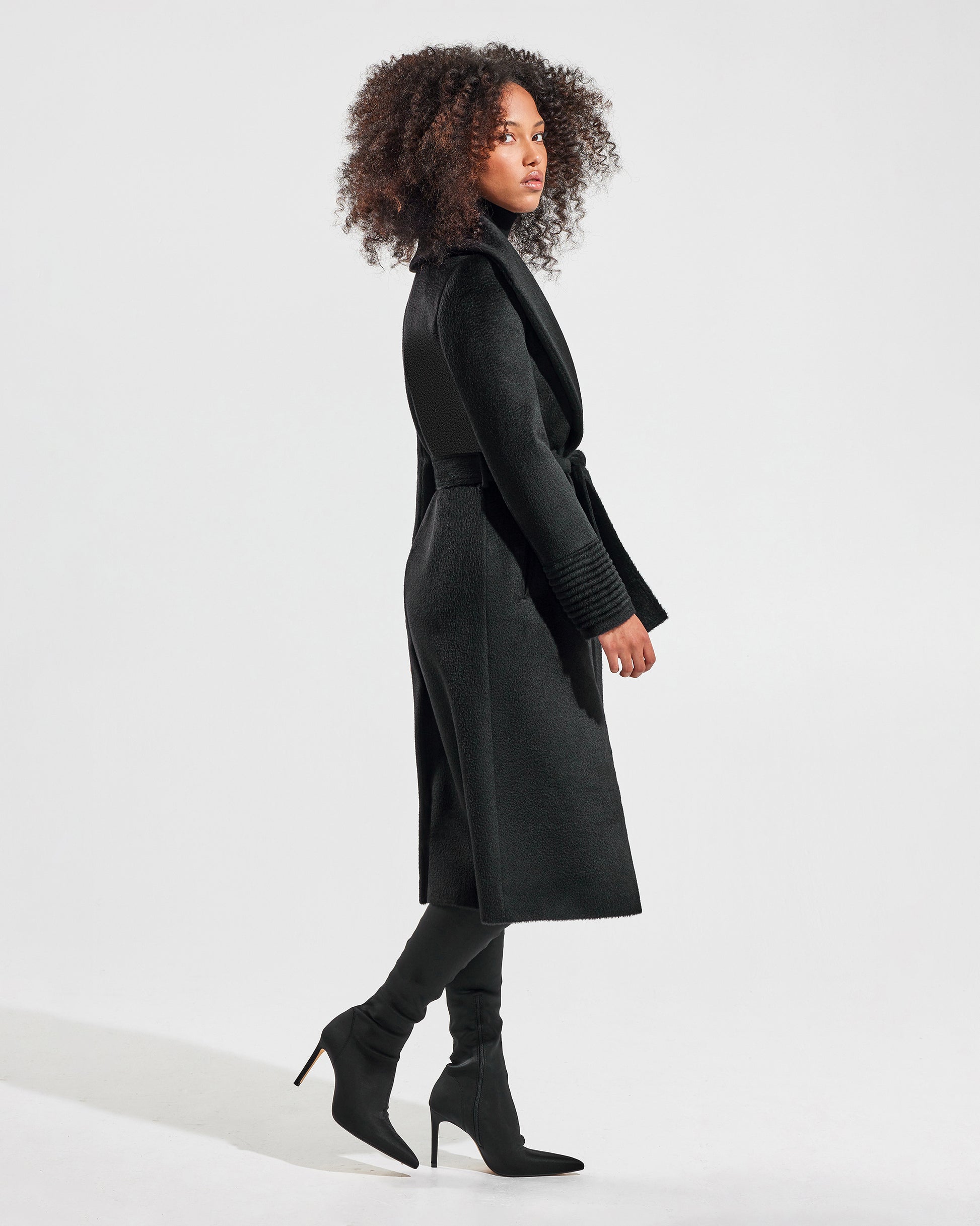 Side view of model wearing a Sentaler long black wrap coat made of suri alpaca wool, with a shawl collar, worn belted.