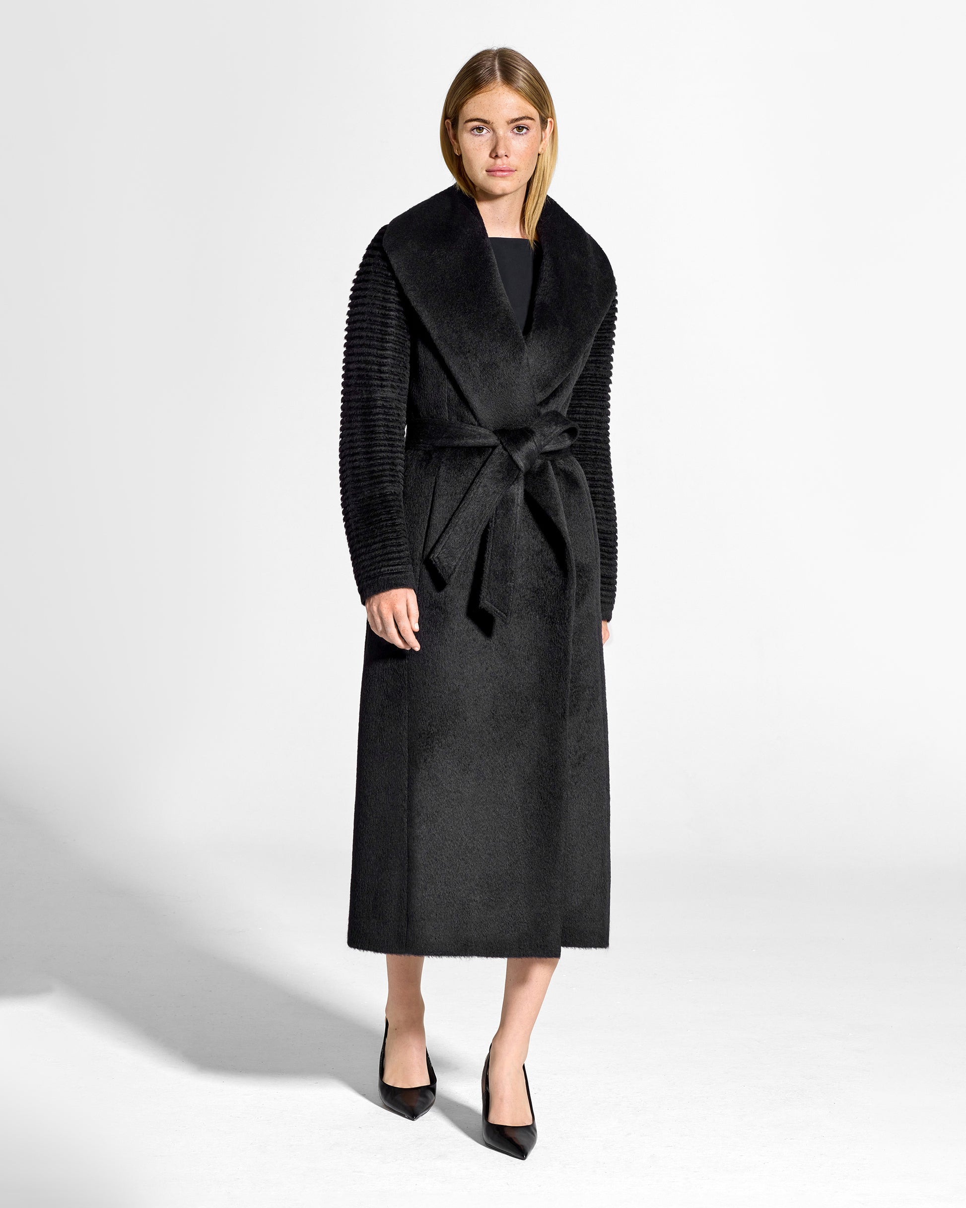 Front view of model wearing a Sentaler long black wrap coat made of suri alpaca wool, with ribbed sleeves, worn belted.