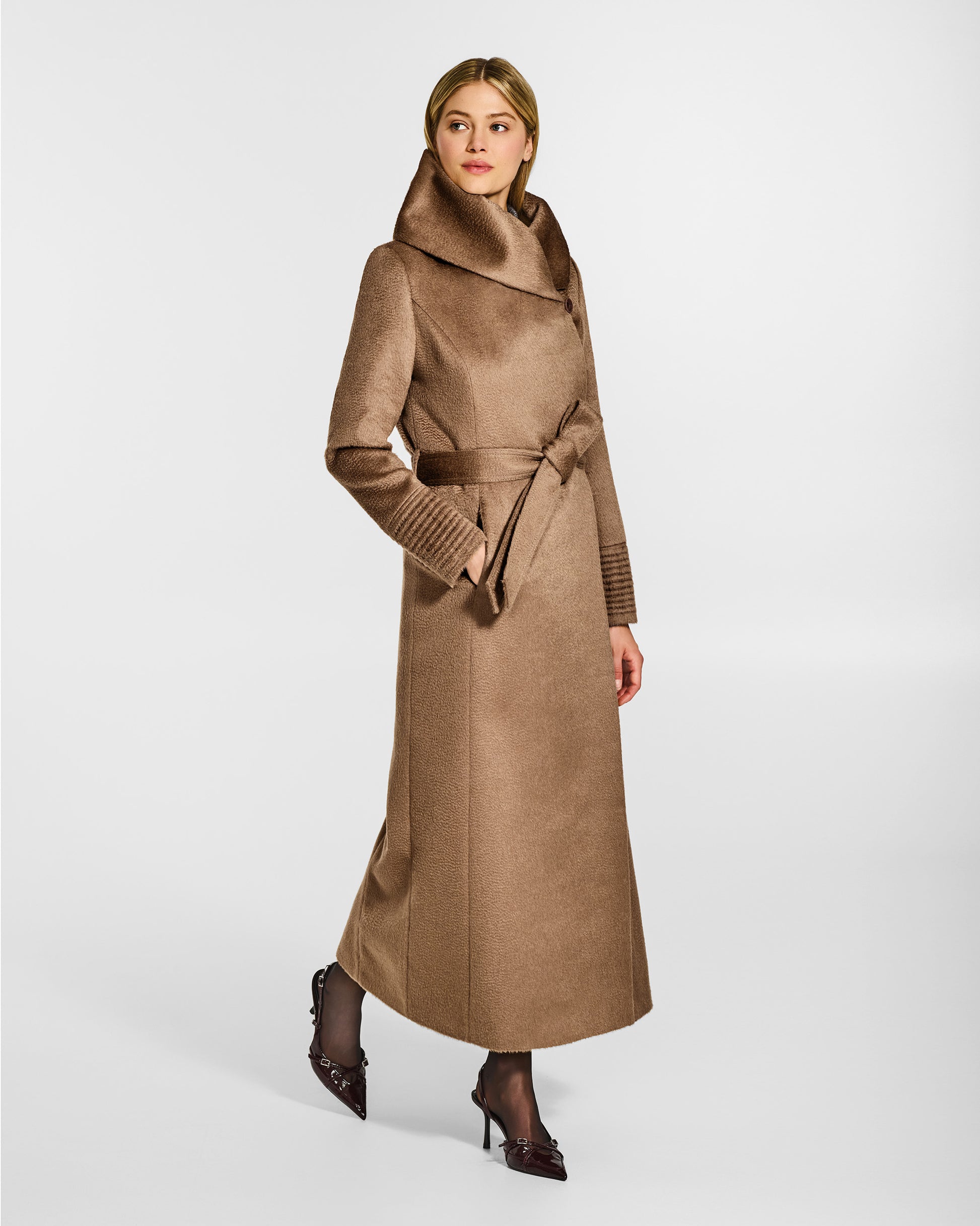 Side view of model wearing a Sentaler maxi dark camel coat made of suri alpaca wool, with a hooded collar, worn belted.