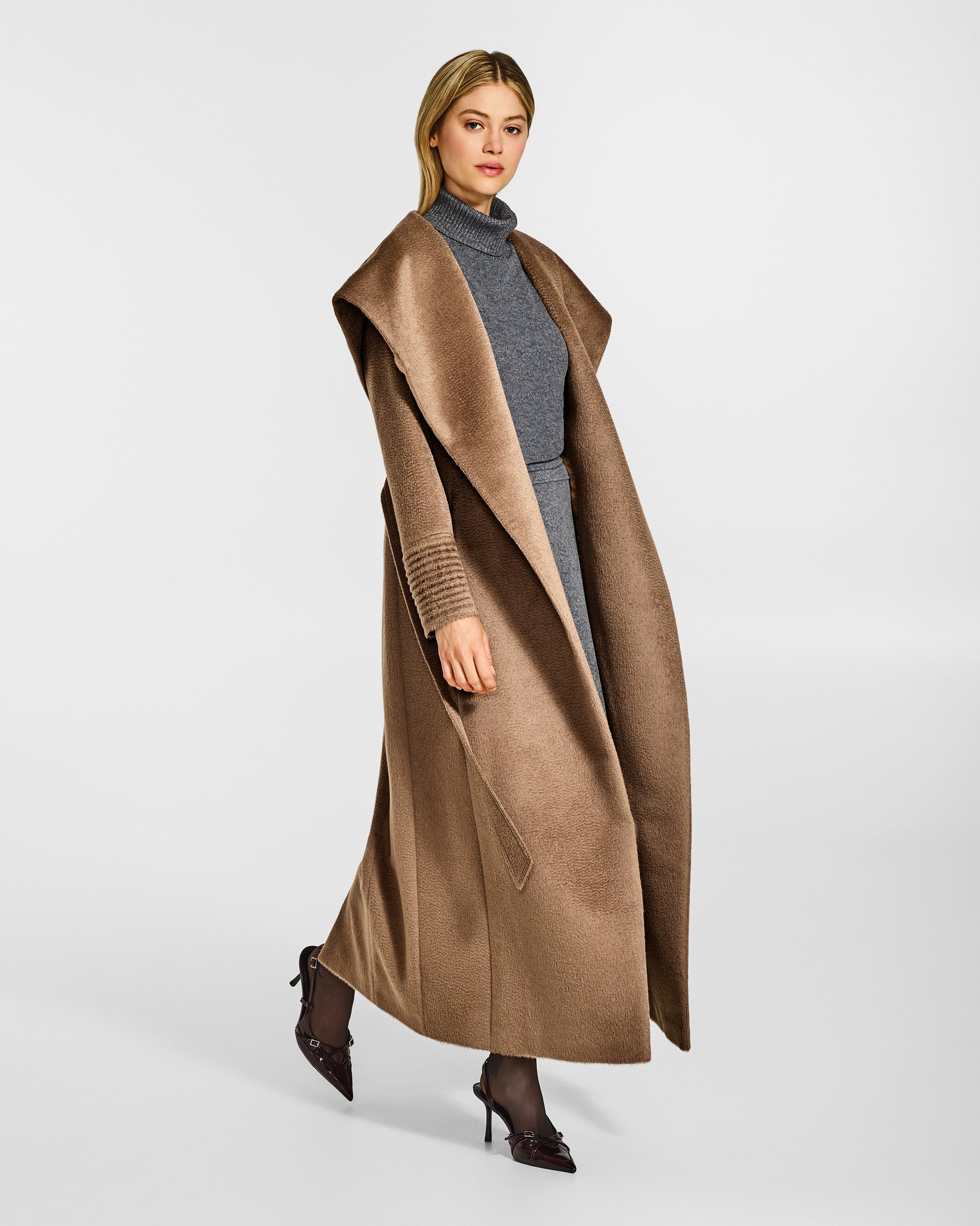 Side view of model wearing a Sentaler maxi dark camel coat made of suri alpaca wool, with a hooded collar, worn open.