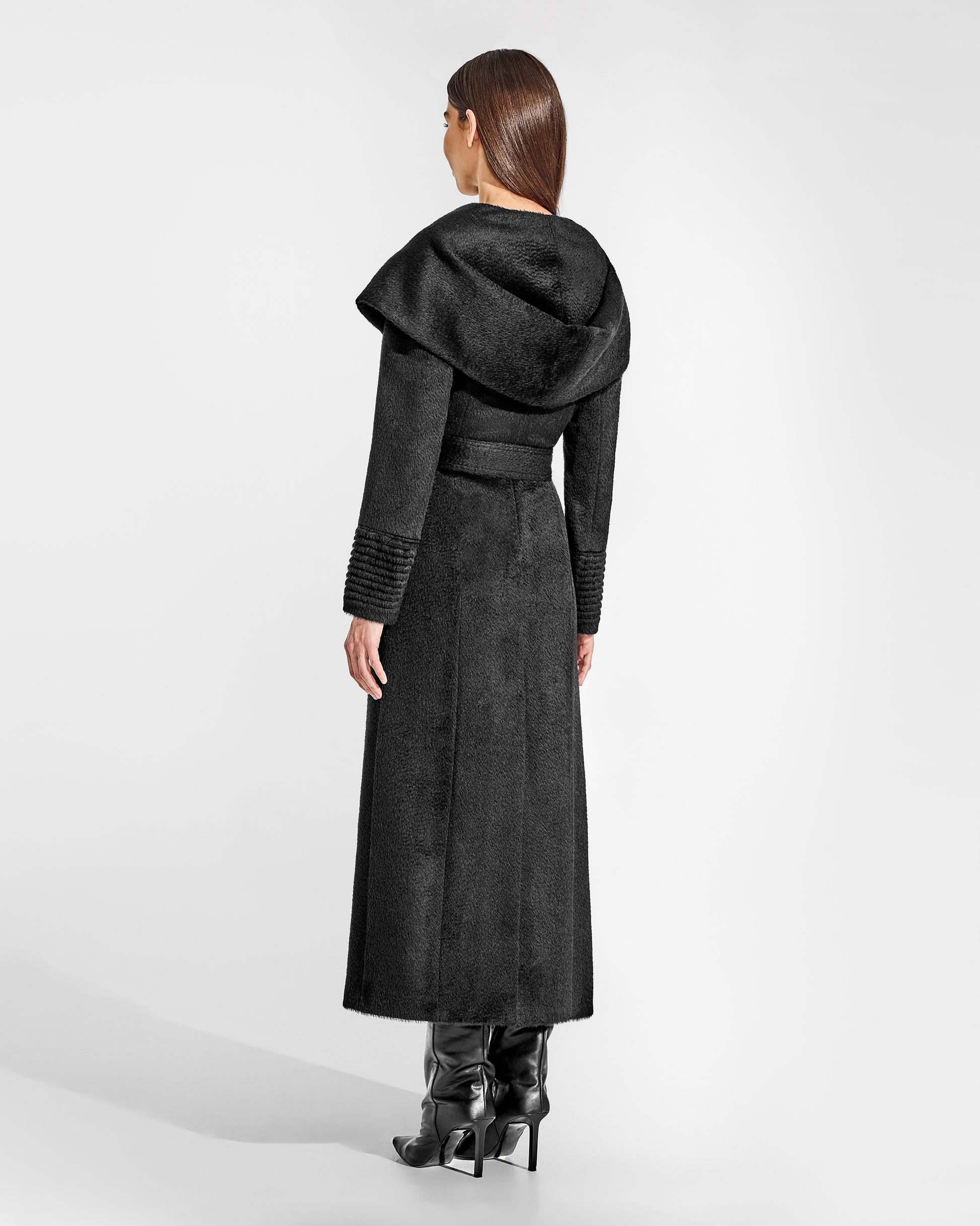 Back view of model wearing a Sentaler maxi black coat made of suri alpaca wool, with a hooded collar, worn belted.