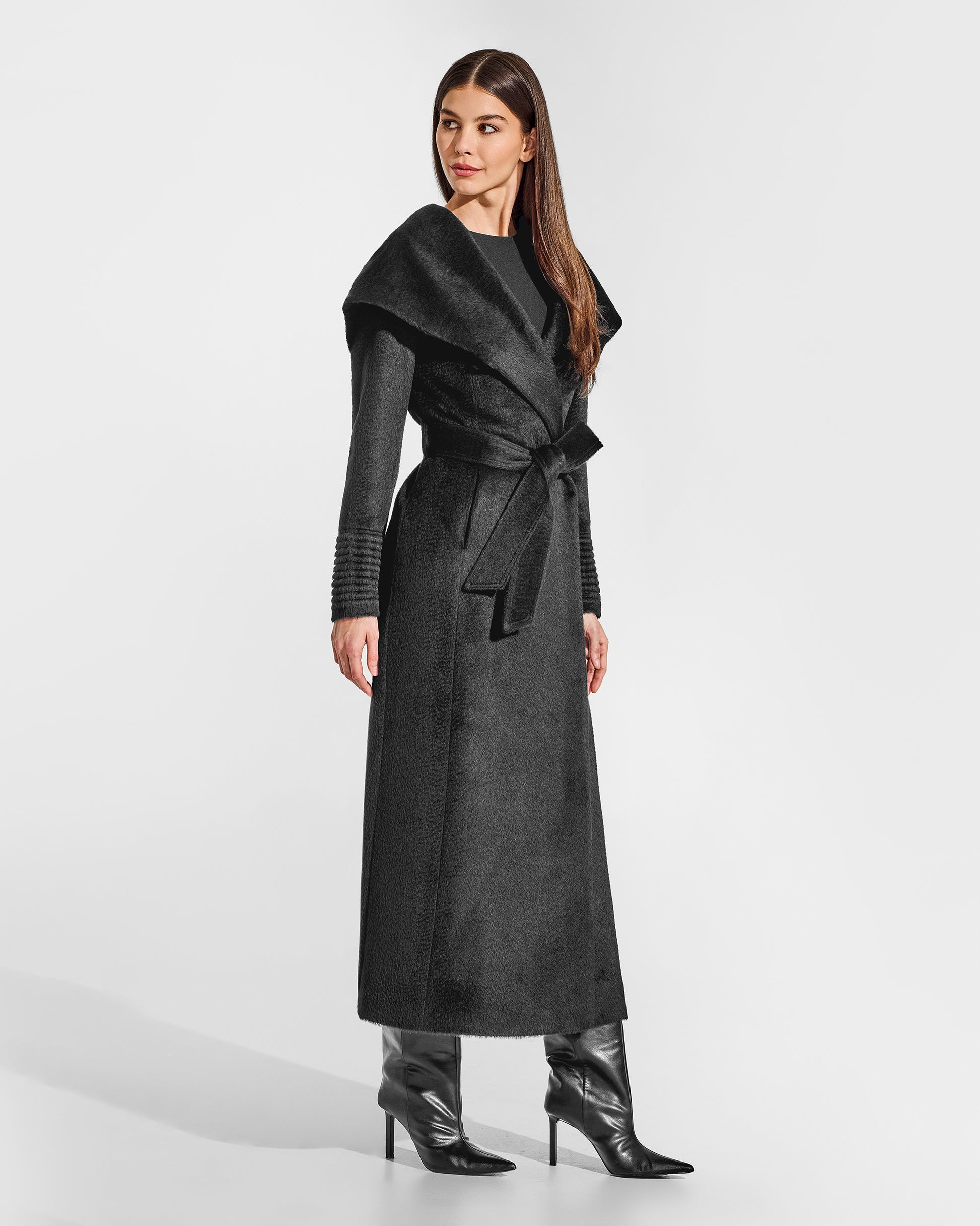 Side view of model wearing a Sentaler maxi black coat made of suri alpaca wool, with a hooded collar, worn belted.