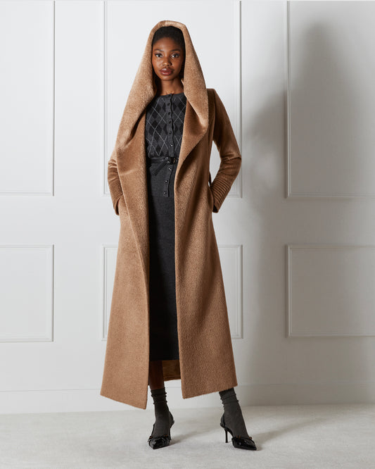 Front view of model wearing a Sentaler maxi dark camel coat made of suri alpaca wool, with a hooded collar, worn open. 