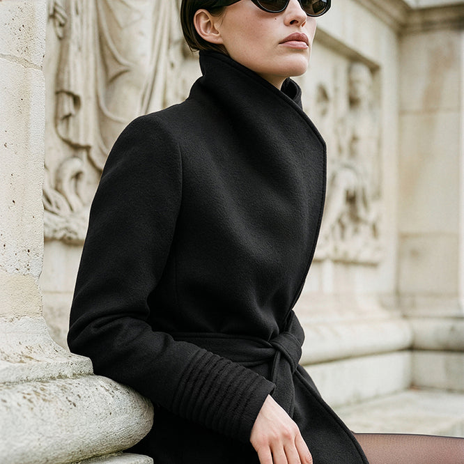 SENTALER Baby Alpaca Long Funnel Collar Wrap Coat in Black, worn by a female model seated against stone architecture.